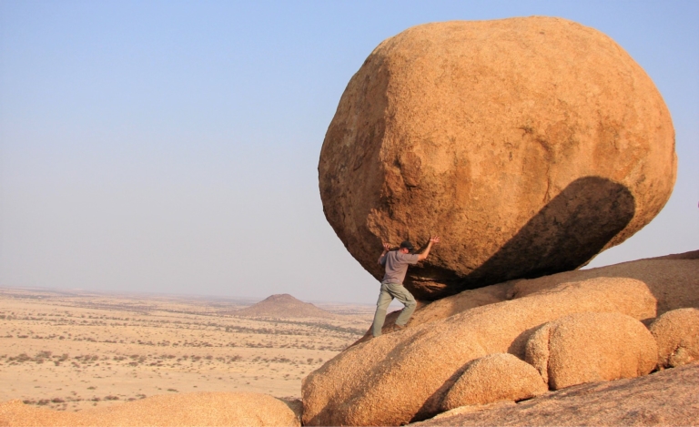 Blues sky with a man pushing a huge boulder up rock ground